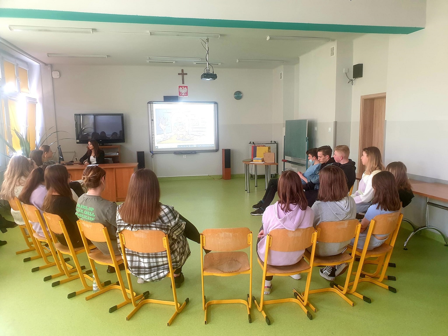 Uczniowie siedzą w klasie w półokręgu, oglądają prezentację na ekranie